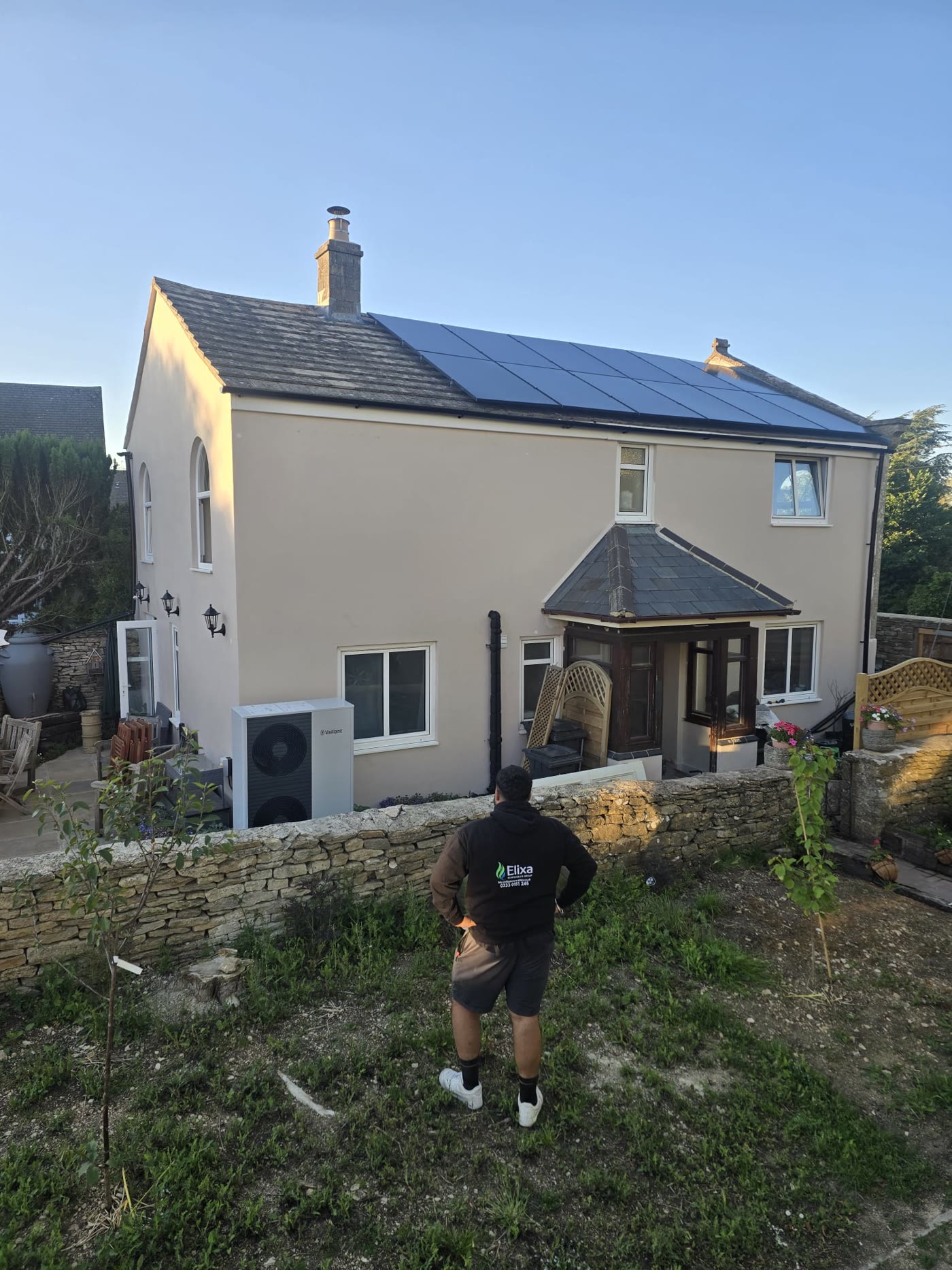 Rendered stone cottage with an all-black solar array and a Vaillant aroTHERM plus heat pump beside the patio, an Elixa engineer surveying from the garden